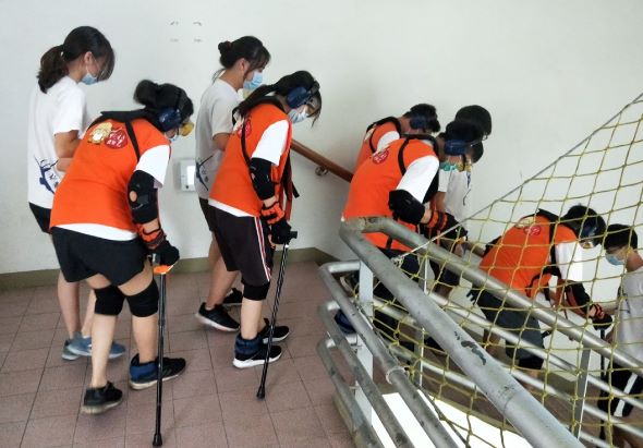 一群穿著橙色運動服的學生在室內樓梯間進行冰球訓練，他們手持冰球棍，站在樓梯旁邊練習動作。教練穿著白色上衣在旁指導，目標架放置在樓梯下方。
