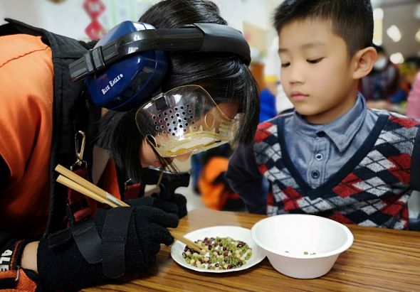 兒童戴著藍色護目鏡和防護面罩進行科學實驗，在木製桌面上觀察盛裝在玻璃容器中的食物樣品，旁邊放著盤子和碗。另一名穿著紅藍色毛衣的男童在旁觀看，背景為室內活動場景。