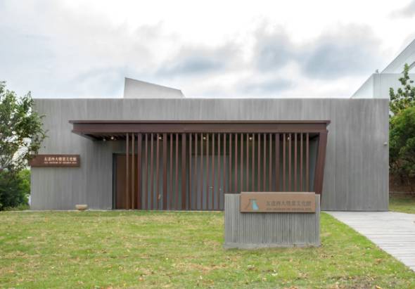 現代建築風格的文化館外觀，灰色金屬波浪板牆面，入口處設有棕色豎條紋柵欄門。建築前有草坪和白色木質步道，左右兩側植有綠樹，陰天背景。