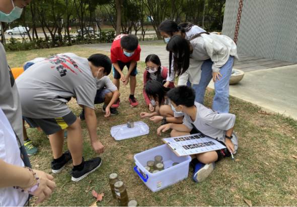 一群學生在戶外草地上圍成一圈，進行自然科學觀察活動。地上放著裝有石頭、貝殼等自然物質的白色容器和紙張記錄，學生們彎身觀察、討論和記錄，背景是公園樹木和建築物。