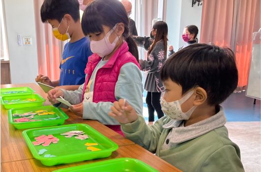 室內課室中，數名戴著口罩的小學生聚集在綠色工作檯前，專注地進行手工藝活動。前景的女童正在操作手中的美勞材料，背景有其他學生和成人在進行相同活動。窗邊有粉紅色窗簾，室內採光充足。