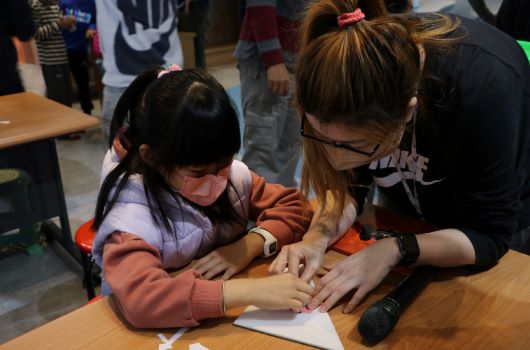 一位戴眼鏡的女性導師協助兩名小女孩在木質桌面上進行手工藝活動。其中一個女孩穿著棕色夾克，另一個穿著紫色上衣。背景有其他參與者和工作人員，環境看起來像是教室或工作坊場景。
