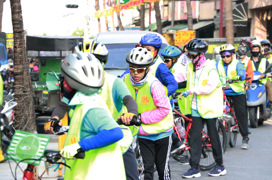 一群穿著螢光背心和安全帽的騎自行車者在城市街道上集合，參加單車活動。他們穿著色彩繽紛的運動服裝，準備出發騎行。背景可見建築物、招牌和其他參與者。