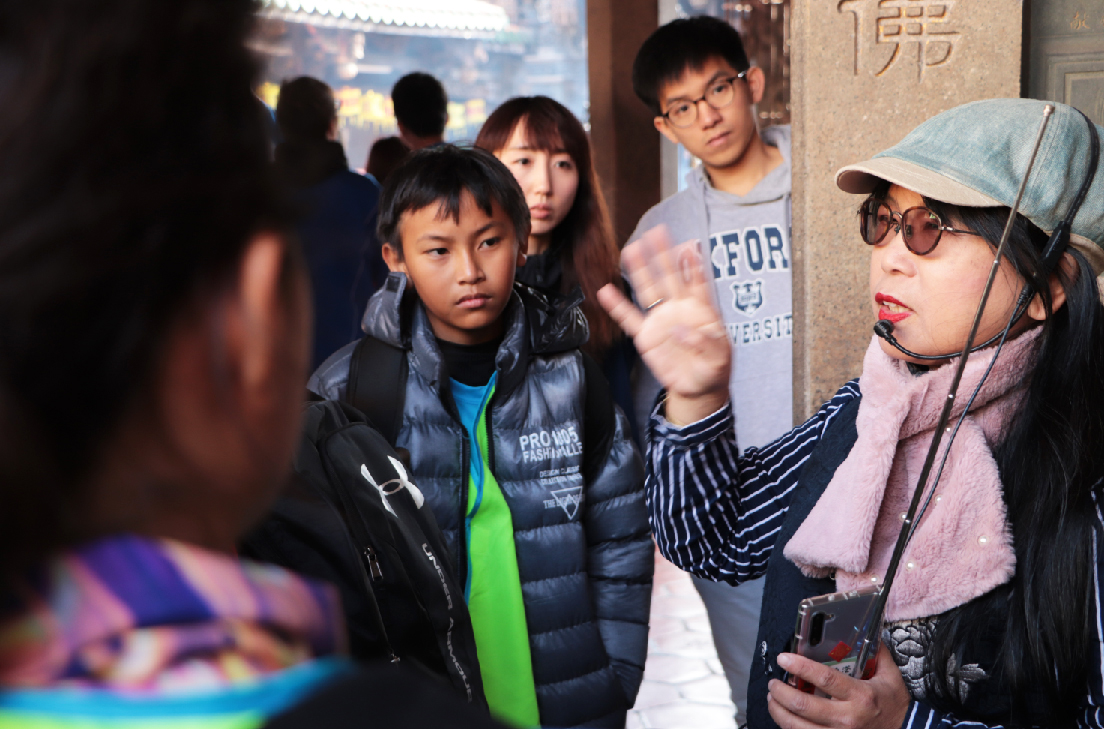 一位戴著鴨舌帽和圓形眼鏡的女性在古色古香的建築前，向一群年輕遊客介紹景點。幾位穿著冬衣的青少年聚精會神地聽講，背景可見傳統中式建築和遊客。