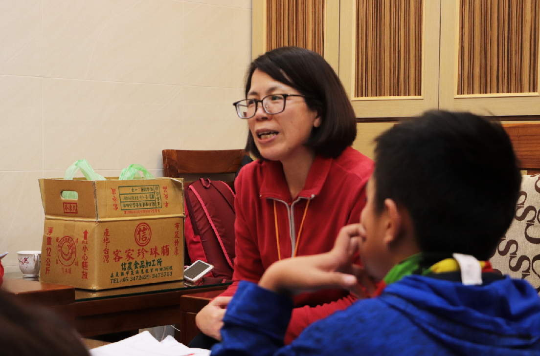 一位穿著紅色外套、戴眼鏡的女性在室內與兒童進行互動,身旁放置著標有中文字樣的紙箱和綠色物品。背景為木製家具和窗簾,氛圍溫暖。