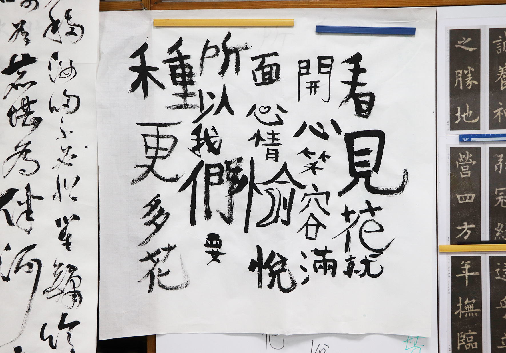 書法練習紙張，中央以楷書書寫一首古詩，筆畫粗重有力。左側為毛筆字練習，右側展示裝裱的古籍書頁。黃、藍色尺規置於上方，展示書法學習的教材與臨帖作品。