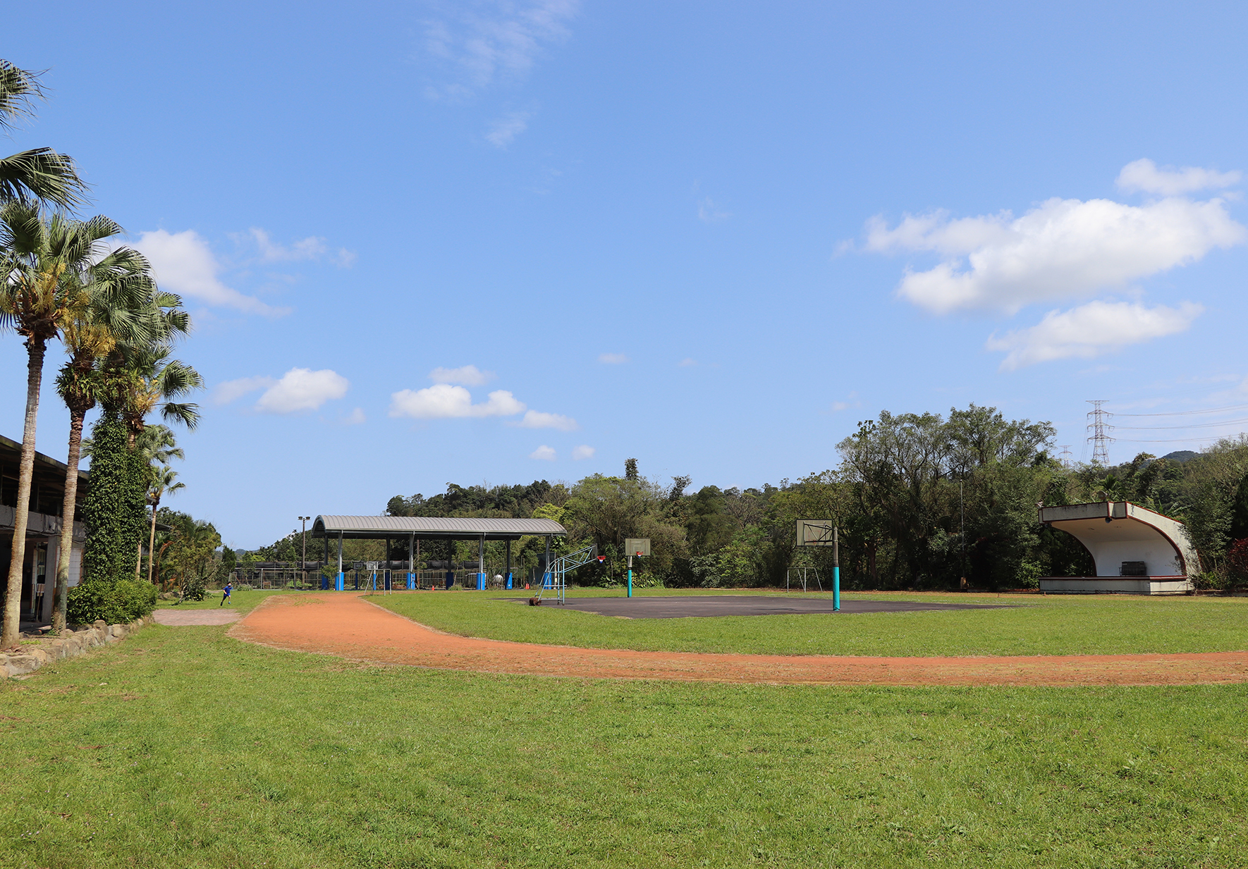 寬敞的社區運動公園，綠色草坪上有紅土棒球場，背景可見遮陽棚、籃球架、半圓形建築物及樹木。藍天白雲，晴朗宜人的戶外休閒環境。