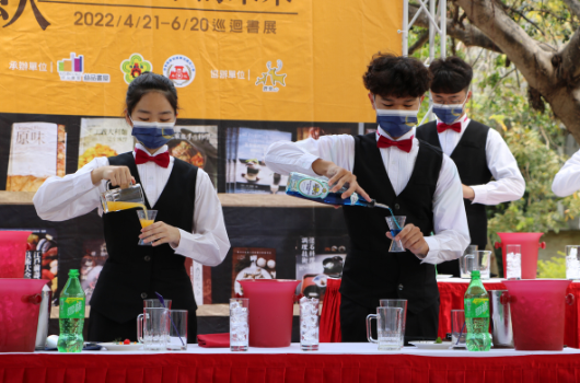 四位穿著正式服裝、戴著口罩的年輕服務員在紅色桌布的長桌前執行調酒表演。背景是2022年4月21至20日的黃色活動看板。桌上擺放玻璃杯、酒瓶和飲料瓶。這是一場專業的餐飲服務或調酒競賽活動。