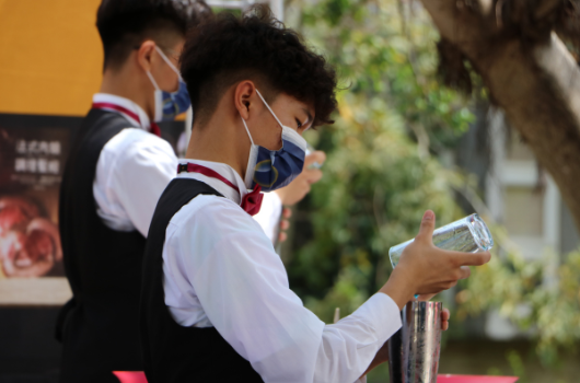 穿著制服、戴著口罩和紅白藍領巾的學生在戶外活動中進行消毒或衛生檢查，手持透明液體容器，身後可見黃色建築物和綠色植物。