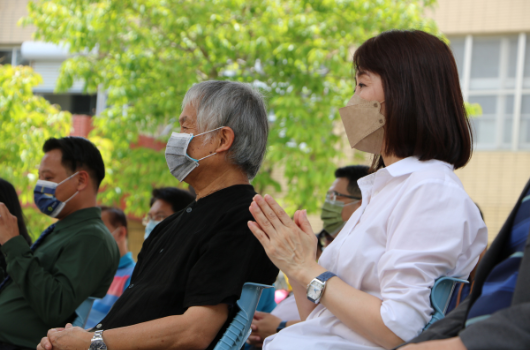 戶外活動現場，多位配戴口罩的參與者聚集，右側穿著白色上衣的女性雙手合十，身旁年長男性穿著黑色衣服，背景為綠色植物和建築物，氛圍溫馨莊重。