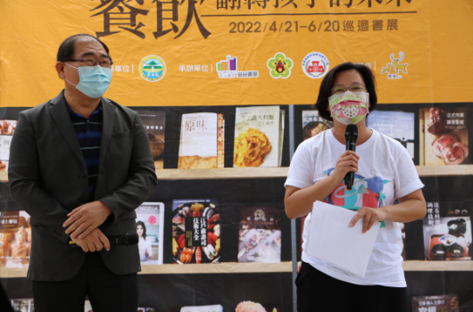 兩位戴著口罩的講者在展覽會現場進行演講。男性穿著黑色外套站在左側，女性穿著白色上衣持麥克風站在右側。身後為黃色背景的展示牆，展示各式食品和書籍。牆面標題為「食飲」，展期為2022年4月21日至6月20日。