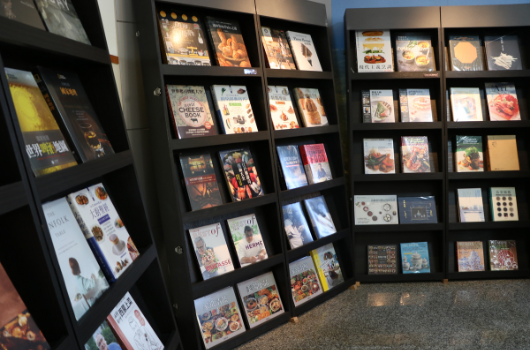 展覽會場展示多個黑色展示櫃，櫃內陳列各式食品包裝、雜誌封面及印刷品等收藏品。左側有電子螢幕播放相關內容，展現設計與文化主題的視覺展覽。