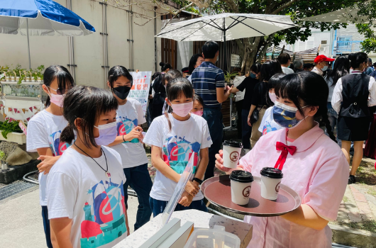戶外校園活動現場，穿著彩色T恤和粉紅色衣服的學生們聚集在帳篷下，參與飲料試飲或販售活動。背景可見其他參與者和綠色植物，天氣晴朗。