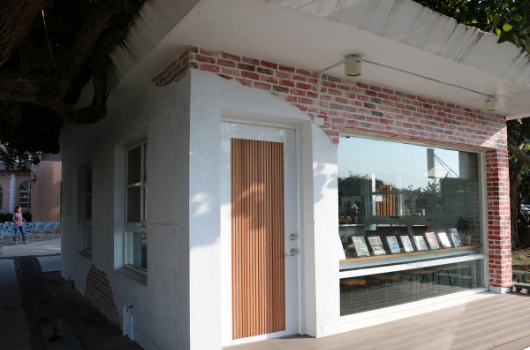 現代建築風格的白色外牆小屋，配備木製豎條紋門和大型玻璃窗。建築物左側為木製柱子，右側為寬敞的木質露台，露台上擺放黑色躺椅。紅磚煙囪和天然採光營造溫暖舒適的居住空間。