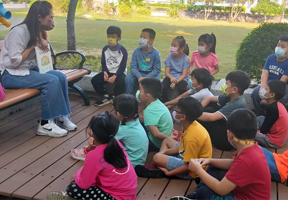 一位女性老師在戶外木製平台上，面對著坐成圓圈的約十五名兒童進行教學。兒童們穿著各色T恤，多數配戴口罩，認真聆聽。背景是綠色草坪和樹木，呈現戶外教學情景。