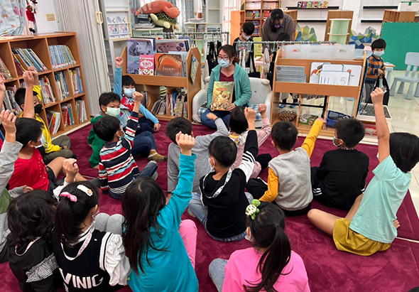 一群小學生坐在圖書館的紅色地毯上，圍成一圈聆聽老師講故事。教室內有書架、展示板和各式教材。老師站在中央手拿書籍，學生們專注地看著，參加閱讀推廣活動。