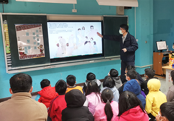 教室裡，一位男性講師站在綠色黑板前指著投影螢幕上的圖片進行教學。螢幕顯示多人合照。約二十位穿著五彩繽紛冬衣的學生坐著聆聽，背對鏡頭面向講台。教室左側黑板上貼有海報。