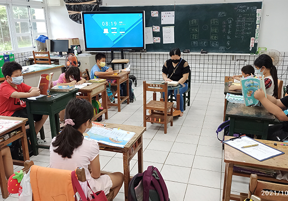 小學教室內，學生戴著口罩坐在木製課桌前進行課程。教師站在講台旁指導學生。黑板上有板書，牆上掛著電子螢幕顯示課程內容。教室採光良好，地板為白色磁磚。