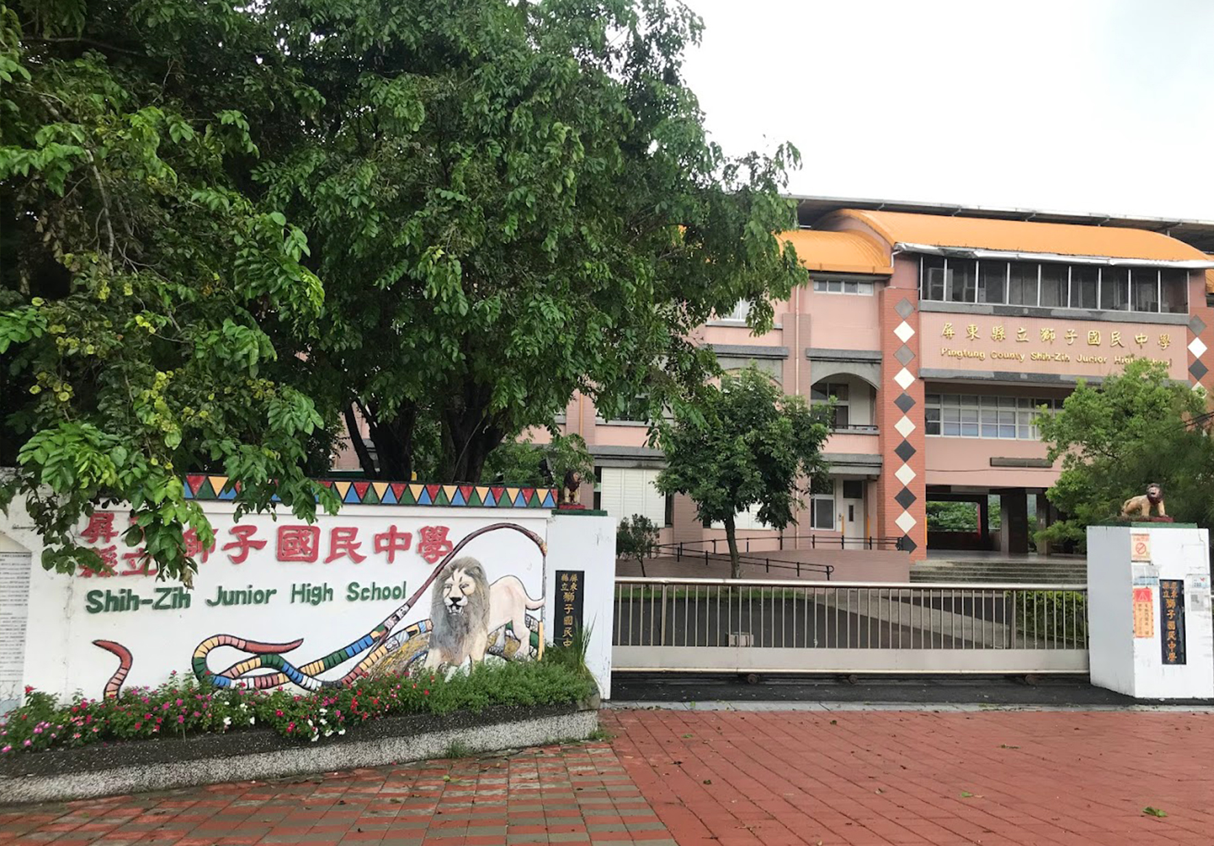 士子國民中學校門外觀，白色圍牆上有校名標誌和傳統獅子圖案裝飾，後方為磚紅色校舍建築，周圍綠樹環繞，校門為黑色鐵門設計。