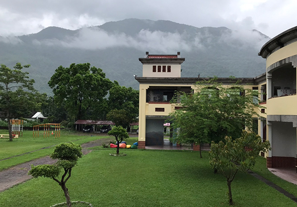 一棟黃色中式建築坐落於綠草坪中，背後聳立著被雲霧籠罩的高山。前院設有兒童遊樂設施、涼亭和小樹，右側是現代建築。遠處山脈雲霧繚繞，營造寧靜的度假氛圍。