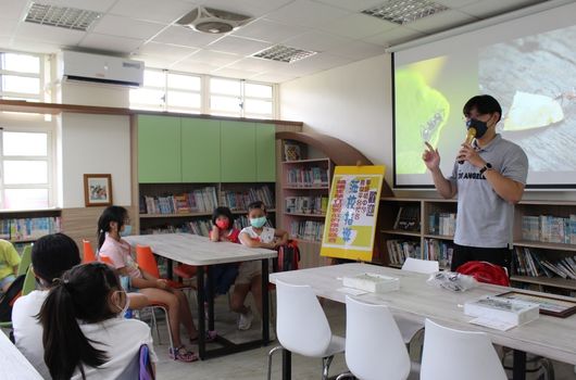 圖書館內，一位講師正向坐在桌前的兒童講授課程。講師站在投影幕前做手勢說明，身旁擺放黃色教學看板。教室內有書架、兒童讀物和教學用具。約五名兒童認真聆聽，參與互動學習活動。