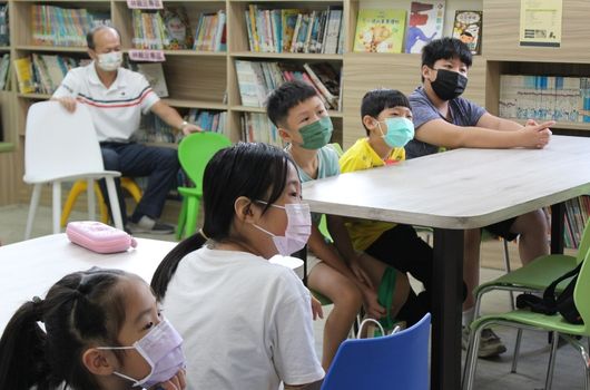 圖書館內,戴著口罩的學生們圍坐在白色桌子前參加閱讀活動。一位成人講師坐在後方,為孩童們進行故事講述或教學。書架上擺放著各式圖書,呈現學習氛圍。