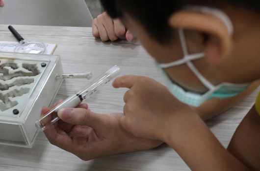兒童戴著口罩在實驗室進行科學實驗，手持吸管對著裝有液體的透明容器進行操作，桌上放有實驗器材和記錄表格，展示STEAM教育學習活動。
