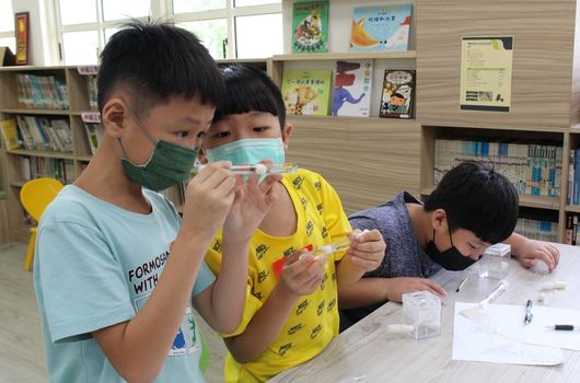 圖書館內，三名戴口罩的小學生進行科學實驗活動。左側兩名學生穿著黃色上衣，正在觀察和操作實驗器材；右側學生穿著深色上衣，專注地進行記錄。背景為書架和教室環境。