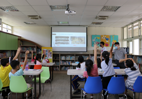 小學教室內，學生穿著黃、藍、紅等多色制服坐在課桌前，面向講台上的投影幕。教師正進行課堂教學，牆上貼有彩色教學海報，後方有書架和教學用具。