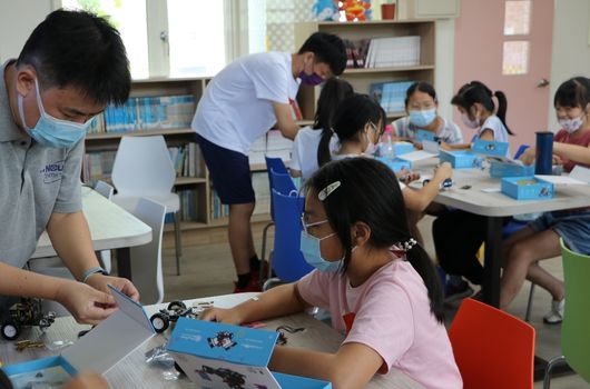 教室內，戴口罩的成人志工與多名小學生進行STEAM教育活動，學生們圍坐在藍色工作盤前組裝機器人或科技教具，專注地進行動手實驗與學習。