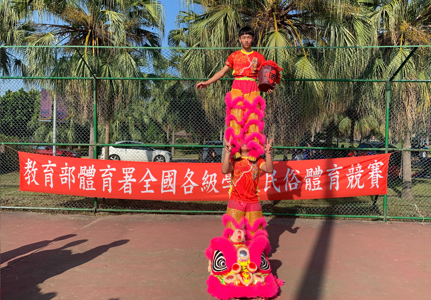 兩名年輕人在室外籃球場進行舞獅表演。前景為粉紅色獅頭，獅身裝飾金色圖案。後方人物穿著紅色中式服裝。背景為綠色圍欄、棕櫚樹與紅色布幅，布幅上有中文字。