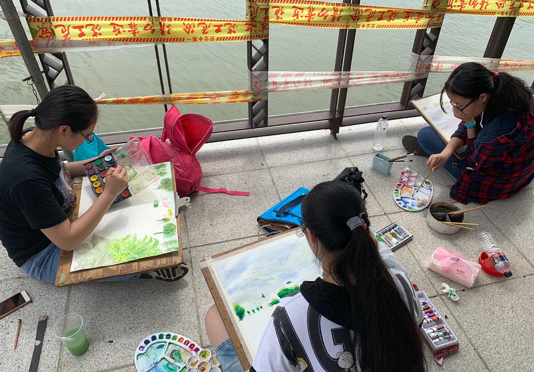 四名學生在靠近河邊的空地上進行戶外繪畫活動。她們各自在木製畫板上作畫，使用水彩顏料和筆刷。周圍散放著畫具、背包和水杯。上方掛著紅黃相間的裝飾帶，背景是河景。
