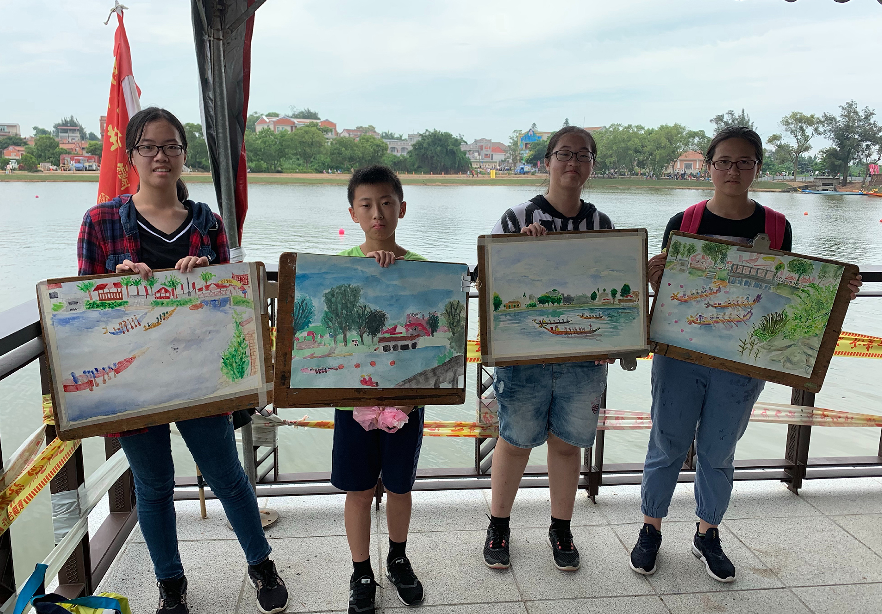 四名學生在河邊展示他們的繪畫作品。背景是寬闊的河流、對岸的建築和紅旗。每位學生各自舉著一幅描繪龍舟競賽、河景和傳統活動的彩色畫作。這是一場展示學生藝術創作的戶外活動。