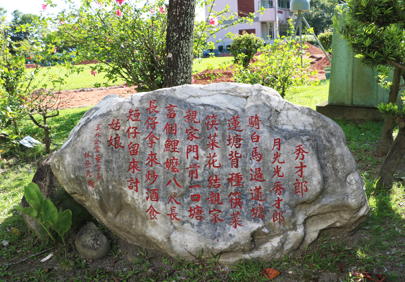 一塊刻有紅色繁體中文詩文的灰色石碑，豎立在綠草地上，周圍環繞著樹木和花卉，背景可見民宅建築。石碑上的文字排列整齊，記載了某段文學作品或歷史紀錄。