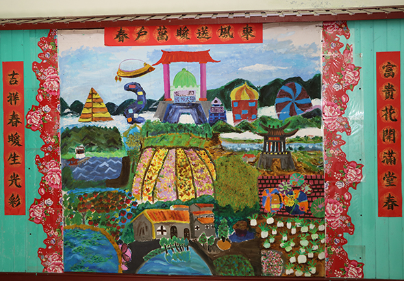 綠色木製板面上繪製的彩色壁畫，描繪農村四季風景。畫面包含稻田、果樹、房屋、水塘、紅色牌樓等元素。四周以紅色裝飾邊框圍繞，框內有金色花卉圖案。左右兩側各有紅色直幅，上方為紅色橫幅，均寫有中文書法字。