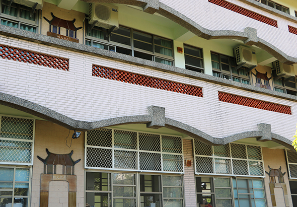 一棟亞洲風格建築，白色磚牆搭配紅褐色瓦片屋頂，上層為黃綠色外牆。建築正面設有多扇綠色鐵窗和玻璃門窗，兩側裝飾著褐色木製飛簷造型。整體展現傳統與現代混合的建築特色。