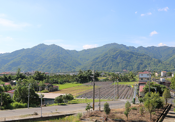 山區農村風景，前景為已開墾和未開墾的田地，中景有稀疏民房和電線桿，背景為層疊翠綠的高山，晴空萬里，呈現寧靜的鄉村田園景觀。