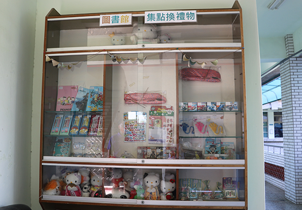 圖書館集點換禮物展示櫃，玻璃櫃內陳列漫畫書、卡通玩偶、文具用品、毛巾等兒童獎品，頂部標示中文字樣，櫃子分為上下兩層，展示各式集點兌換禮物。