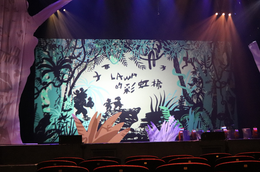 劇院舞台上展示森林主題的投影畫面，描繪樹木、植物和動物剪影，舞台下方有紫色燈光照射的樂器和設備，觀眾座位在前景，現場進行表演或演唱會活動。