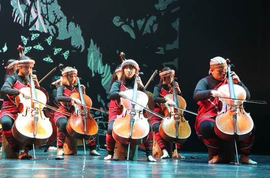 五位音樂家身穿紅藍相間服裝，在舞台上演奏大提琴。舞台背景為深色投影幕，展示地球地圖。演奏者戴著白色帽子，專注於演奏樂器，呈現古典音樂表演的專業舞台場景。