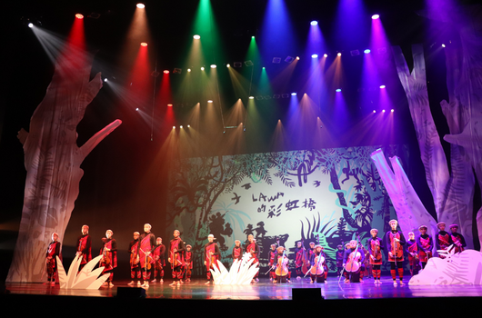 舞台上約20位演員穿著紅色服裝排成一列，背景為大型投影屏幕展示傳統風景圖案。舞台兩側裝置白色和粉紅色蓮花造型燈飾，頭頂彩色聚光燈照亮整個舞台，呈現盛大的文藝表演場景。
