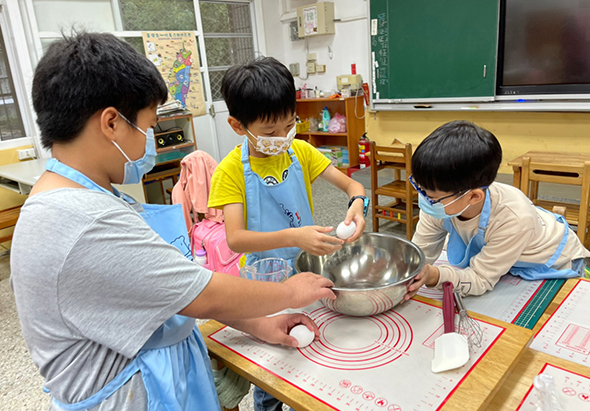 三名戴著口罩的小學生在教室內進行烹飪課程，圍著一張鋪著紅色墊子的桌子。左側學生穿著藍色圍裙，中間學生穿黃色上衣，右側學生穿米色上衣，三人正在金屬碗中攪拌食材。桌上放著白色盤子、擀麵杖等烹飪用具。