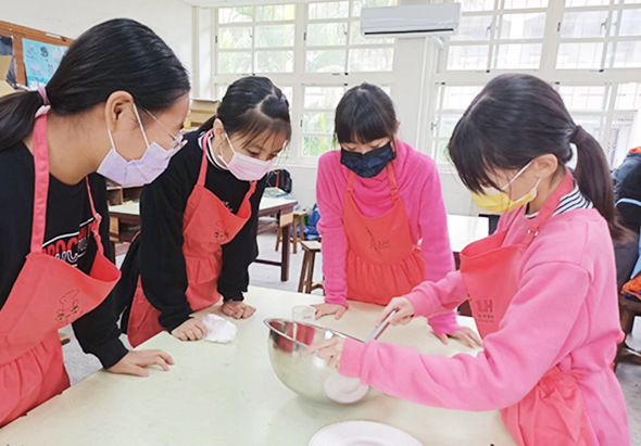 五位戴著口罩、穿著紅色圍裙的學生在教室內圍繞白色工作台進行烹飪或食品製作活動，其中一位學生正在攪拌碗中的食材，其他學生專注地觀看或協助，背景可見大型窗戶。