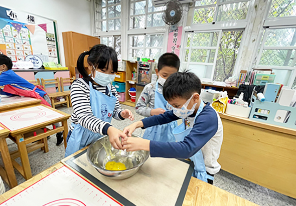 幼兒園教室內，四名戴著口罩的小朋友圍在桌邊，正在進行烹飪活動。他們一起將食材放入金屬碗中，共同參與料理過程。教室背景有大窗戶、布告欄和教學用具。