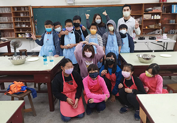 學校烹飪課程中，學生們穿著圍裙和口罩，分成兩組在教室內合影。前排四位學生蹲姿擺pose，後排約十位學生站立，教室背景可見黑板、書架及烹飪用具。
