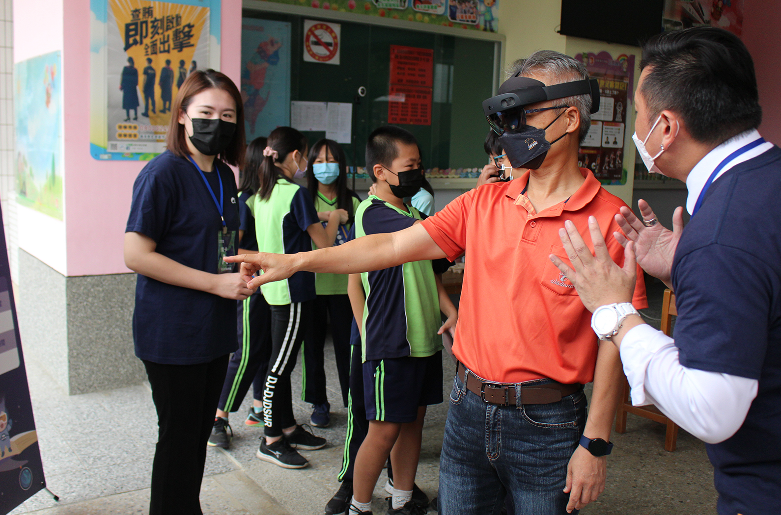 學校教室內，一位穿著紅色polo衫的成人與多位穿著綠黑色運動服的學生進行互動。成人戴著黑色口罩和VR頭盔，與學生進行握手或團隊合作的活動。現場有多名穿著藍色制服的工作人員和穿著深藍色西裝的訪客在場。背景牆上貼有中文海報和教學資料。