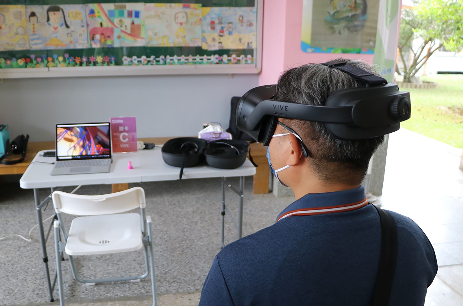 一名戴著HTC Vive虛擬實境頭盔的人在教室內體驗VR應用。桌上放著筆記型電腦，顯示VR內容畫面。背景牆面貼滿學生的彩色美術作品。窗外可見校園綠地。