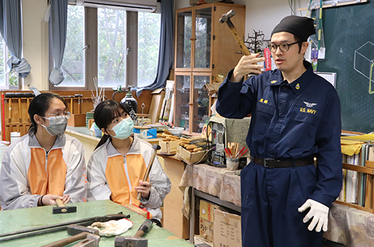木工教室內，一位穿著深藍色工作服的講師向兩位穿著橙色背心、戴著口罩的學生示範木工工具的使用方法。教室裡擺放著各種木工工具和材料，窗戶外可見樹木。