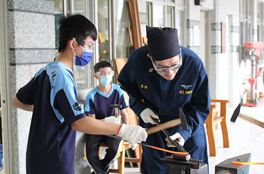 美國海軍人員在室內進行工作培訓，右側軍人穿著深藍色制服戴眼鏡，正使用工具進行操作演示。左側人員穿著黑藍相間制服戴口罩觀看學習。背景有其他參與者，室內設有工作台和各類工具。