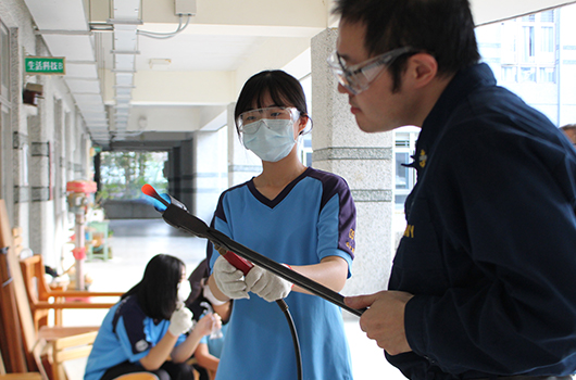 學校走廊內，一名戴口罩的女學生與一位男性教師查看樂器。教師手持木管樂器向學生講解，另有學生坐在地上。背景可見綠色逃生標誌和教室門窗。